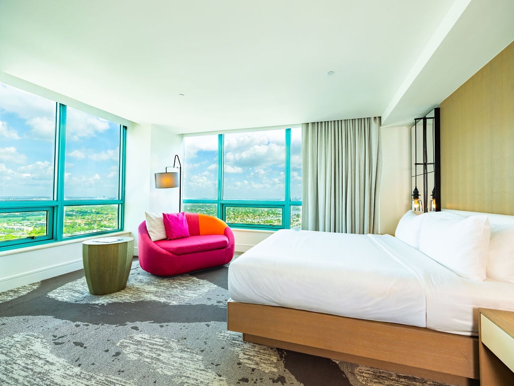 A modern hotel room features a white bed and a vibrant pink and orange sofa by expansive turquoise-framed windows, offering a scenic view of a city landscape.