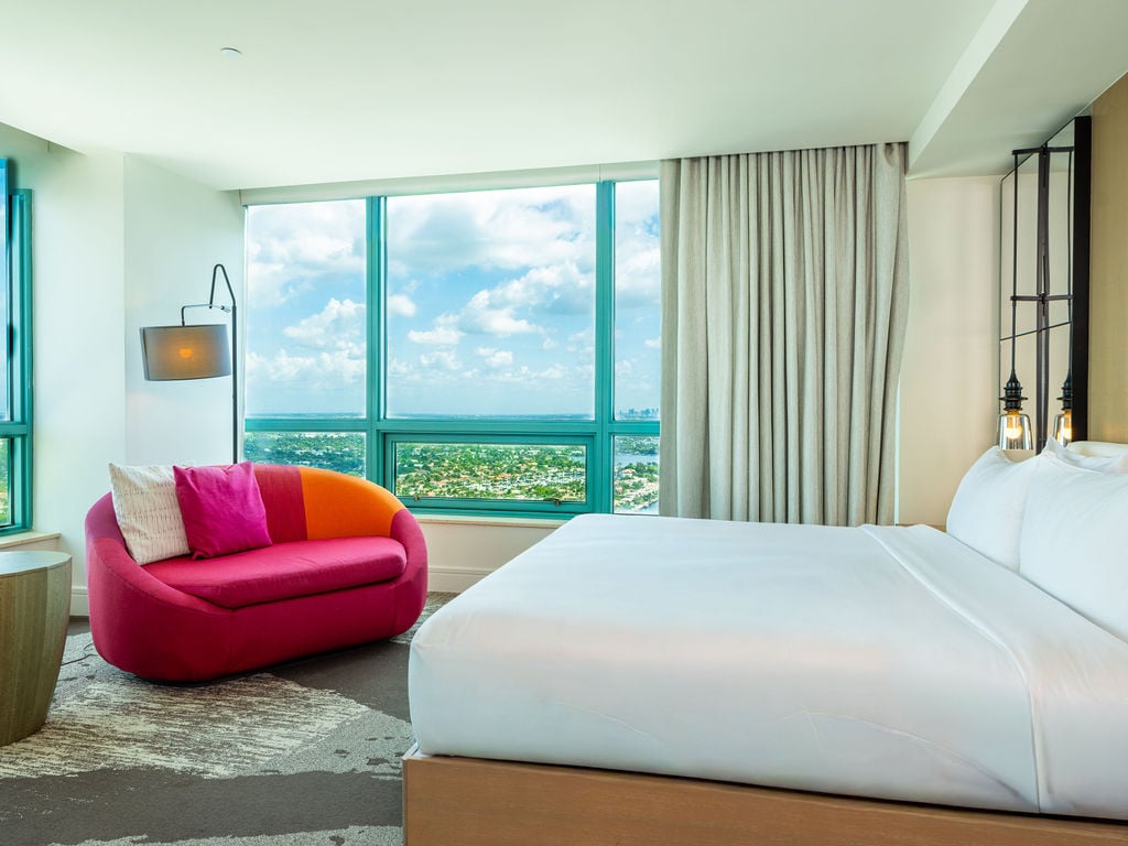 A modern hotel bedroom features a white bed and a vibrant pink sofa, positioned by large windows offering a high-rise view of a city and bay under a cloudy sky.