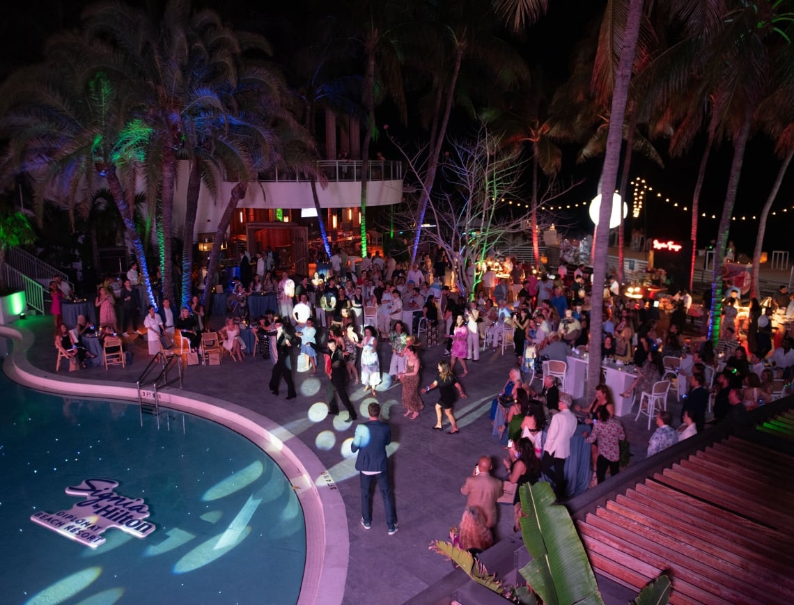 An outdoor evening resort event by a pool at night, featuring a crowded patio, palm trees, and colorful lighting.