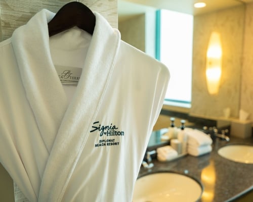 A white Signia by Hilton plush robe hanging on a wooden hanger in a resort bathroom next to a lit vanity mirror.