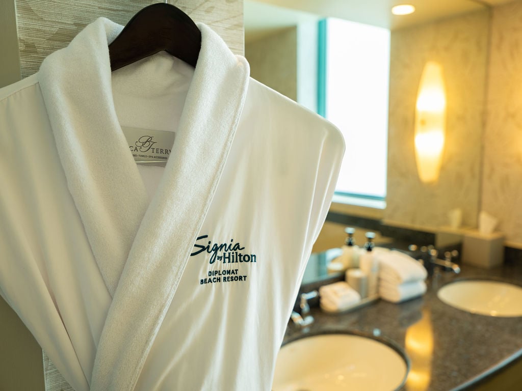 A white Signia by Hilton plush robe hanging on a wooden hanger in a resort bathroom next to a lit vanity mirror.