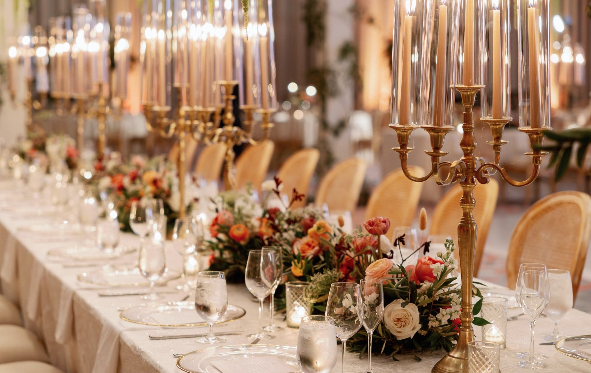 A long dining table is elegantly set for a formal event, featuring lit gold candelabras, vibrant floral centerpieces, and place settings with clear plates and glassware, all in a warm indoor venue.